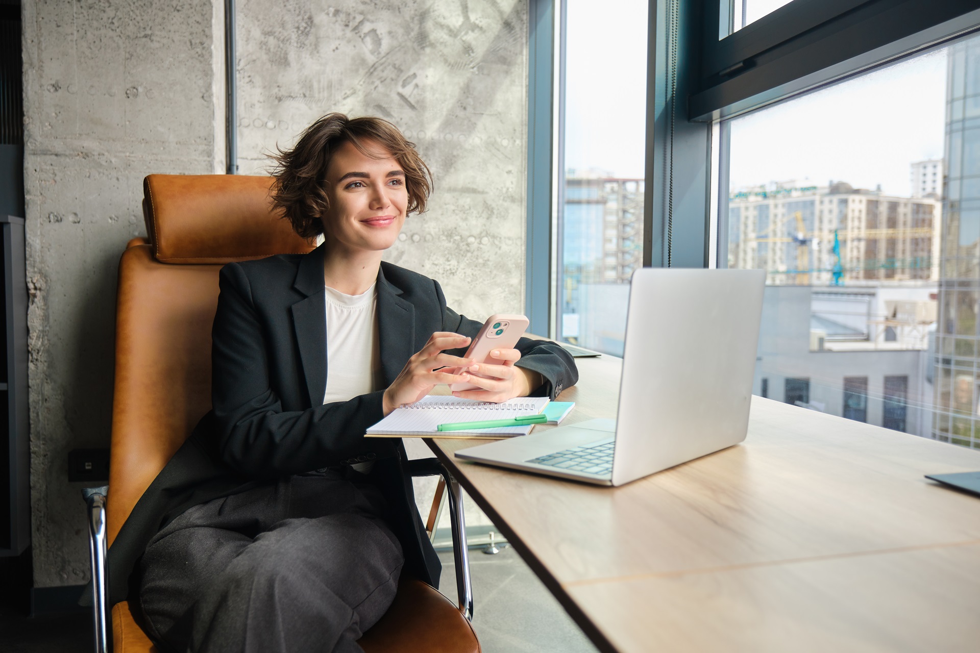 Image of young saleswoman in suit, working on laptop, sitting in her office with smartphone, answering message, reading on mobile phone, using application